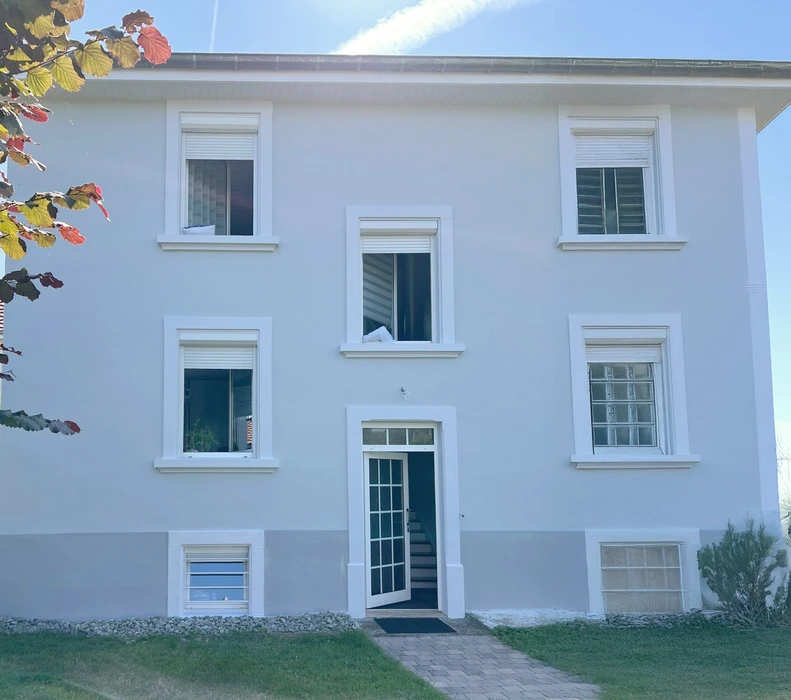 Façade d'une maison résidentielle moderne avec étage, murs enduits beige et gris, fenêtres à encadrements foncés, toiture tuiles terre cuite